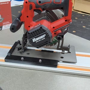 May include: A red and black Bauer 20V 7 1/4" circular saw with a black base is shown on a gray and orange track. A red and white box with the saw's image is in the background. The saw is cordless and brushless.