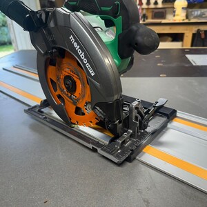 May include: A Metabo HPT circular saw, featuring a black and green body with an orange blade, is positioned on a silver track with orange stripes. The saw is designed for precision cutting in woodworking and construction projects.
