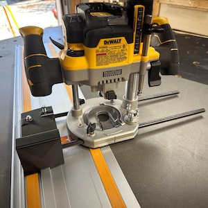 Puede incluir: Una fresadora DeWalt DCW600 amarilla y negra montada sobre una mesa. La fresadora tiene una base plateada y asas con detalles amarillos. La mesa tiene una superficie negra con un riel guía naranja y plateado.