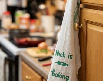 May include: A white kitchen towel with green stripes and the text "Nick is Baking" in green script. The towel hangs from a gold cabinet knob in a kitchen setting.