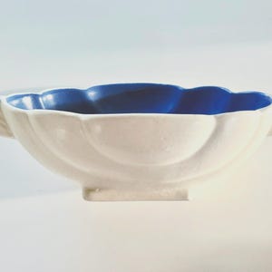 May include: A white ceramic bowl with a scalloped rim and a vibrant blue interior. The bowl has two handles and a rectangular base. The design is reminiscent of a flower, making it a decorative piece for serving or display.