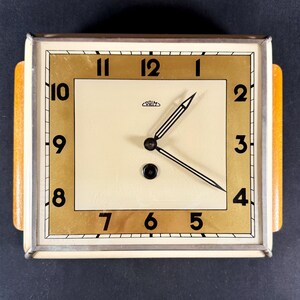 May include: A vintage square clock with a cream-colored face and gold-toned border. Black numerals mark the hours, and the clock hands are black with white stripes. The clock has a wooden frame with a light brown finish.