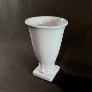 May include: A light blue ceramic vase with a square base and a flared top. The vase has a smooth, glossy finish and a decorative floral design near the base. The vase is approximately 6 inches tall.