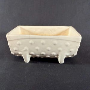 May include: A small, off-white rectangular ceramic planter with a textured, dotted exterior. The planter has three small feet and a smooth interior. The overall design is simple and elegant, suitable for small plants or succulents.