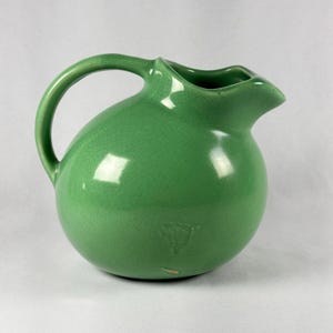 May include: A green ceramic pitcher with a rounded body, a curved handle, and a spout. The pitcher has a glossy finish with a crackle effect. The pitcher is a light green color.