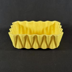 May include: A yellow rectangular ceramic dish with a zig-zag edge and geometric design. The dish is a light yellow color and has a textured surface. The dish is empty and sits on a black surface.