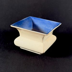 May include: A square, off-white ceramic dish with a flared base and a bright blue interior. The dish has a slightly aged appearance, with subtle variations in the glaze. The design is simple and elegant, suitable for various decorative or functional uses.