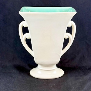 May include: A cream-colored ceramic vase featuring a square-shaped opening with a bright turquoise interior. The vessel has a pedestal base and two symmetrical, curved handles on either side, extending from the upper rim to the lower body.