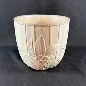 May include: A cream-colored ceramic planter with a floral relief design. The planter has vertical ribbed detailing and a rounded shape. The floral design features leaves and berries. The interior is a lighter cream color.