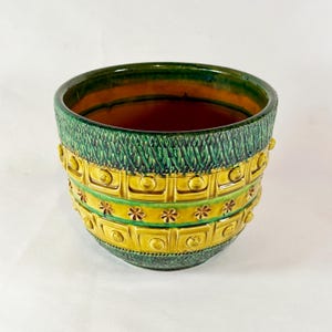May include: A ceramic planter with a textured green and yellow design. The top and bottom sections are green, while the middle section is yellow with embossed squares and floral accents. The interior is a warm brown color.