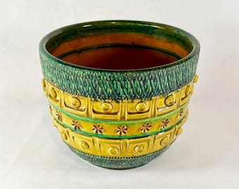 May include: A ceramic planter with a textured green and yellow design. The top and bottom sections are green, while the middle section is yellow with embossed squares and floral accents. The interior is a warm brown color.