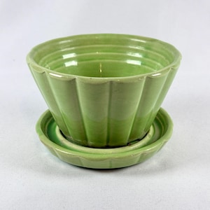 May include: A light green ceramic planter with a fluted, scalloped exterior design and a matching circular saucer base. The bowl features horizontal ridges near the rim and vertical panels extending down to the base, finished in a glossy glaze.