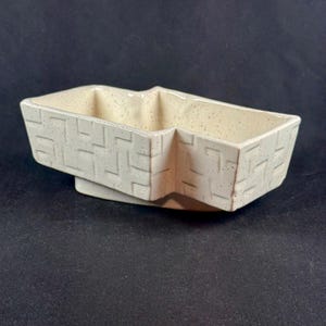May include: A rectangular ceramic dish with a cream-colored speckled glaze and a raised geometric basket-weave pattern on the exterior walls. The vessel features a wide, open top and a sturdy base, presenting a uniform textured finish throughout.