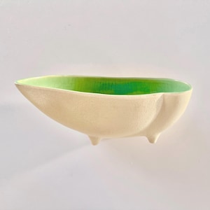 May include: A cream-colored ceramic bowl with a vibrant green interior. The bowl has a unique, elongated shape and rests on three small, rounded feet. The interior color transitions from a bright green to a darker shade, creating a striking contrast.