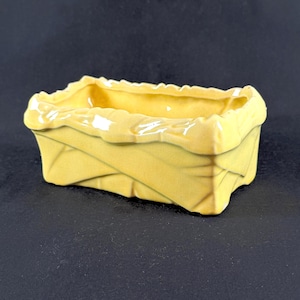 May include: A small, rectangular yellow ceramic planter with a woven basket design. The planter has a ruffled edge and is set against a dark background. The ceramic is glossy.