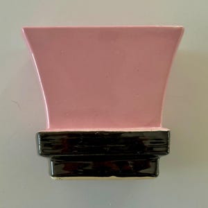 May include: A pink ceramic vase with a flared top and a black base. The vase has a glossy finish and a rectangular shape. The design is simple and modern, suitable for displaying flowers or as a decorative piece.