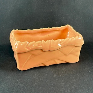 May include: A peach-colored ceramic planter with a basket weave design. The rectangular planter has a ruffled top edge. The interior is smooth, and the exterior has a textured, woven pattern. The planter is suitable for small plants or succulents.