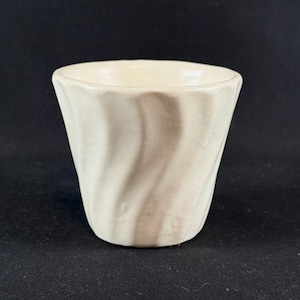 May include: A small, off-white ceramic cup featuring a distinct vertical spiral or fluted pattern along its exterior surface. The vessel has a wide, open circular rim and a tapered base, showcasing a smooth, glazed finish throughout the exterior.