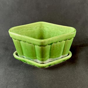 May include: A square, light green ceramic planter with a matching saucer. The planter features a scalloped top edge and vertical fluted sides. The glaze has a glossy finish with visible signs of wear and aging along the bottom base edges.