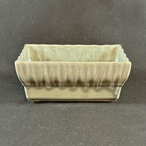 May include: A rectangular ceramic planter with a light green and brown glaze. The planter has a textured, ridged design around the top edge and a smooth base. The glaze has a dripped effect, adding visual interest.