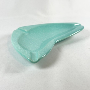 May include: A light turquoise ceramic ashtray with a glossy finish and a speckled texture. The ashtray features an elongated, asymmetrical, kidney-bean shape with three distinct notches along the rim for resting items, showcasing a smooth surface.