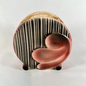 May include: A ceramic napkin holder with a rounded shape, featuring a pink and black color scheme. The front has vertical black and white stripes, and a pink, heart-shaped design. The holder rests on three small black feet.