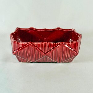 May include: A rectangular, ruby-red ceramic planter with a textured, geometric design. The planter has a scalloped rim and vertical lines that create a diamond pattern on the sides. The interior is a glossy, darker red.
