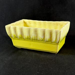 May include: A rectangular, yellow ceramic planter with a ribbed upper edge and a smooth, solid base. The planter has a slightly glossy finish and is set against a dark background. The design is simple and classic, suitable for small plants or succulents.