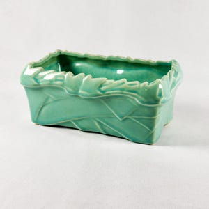 May include: A rectangular, turquoise ceramic planter with a leaf design. The planter has a textured surface with leaf vein details and a scalloped rim. The interior is smooth and glossy, and the planter sits on a white surface.