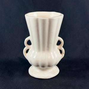May include: A cream-colored ceramic vase featuring a fluted, vertical ribbed design on the upper body and a rounded, bulbous base. The vessel includes two symmetrical, double-loop handles on each side and rests on a circular pedestal foot.