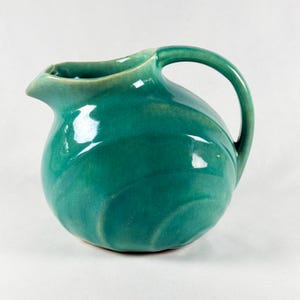 May include: A teal-colored ceramic pitcher with a rounded body and a curved handle. The pitcher has a spout for pouring and a glossy finish. The design features subtle, swirling patterns on the surface.