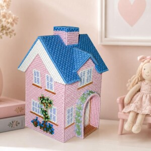 May include: A pink decorative house with a blue roof, white trim, and a chimney. The house features windows, an arched doorway, and floral accents. The house is made of a woven material and sits on a white surface.