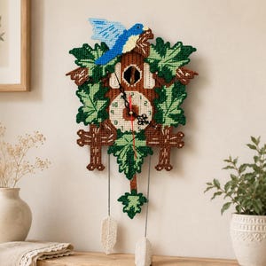 Puede incluir: Un reloj de cuco artesanal con un pájaro azul en la parte superior. El reloj presenta un marco de madera marrón, detalles de hojas verdes y una esfera crema con números negros y manecillas rojas. El reloj cuelga de dos pesas color crema.