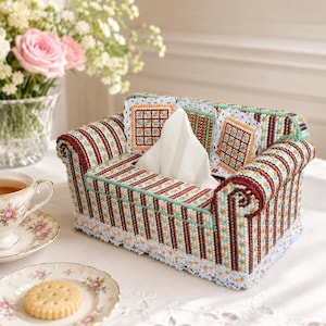 Könnte beinhalten: Dekorative Taschentuchbox in Form eines Miniatursofas. Die Box hat ein gestreiftes Muster in Burgunderrot, Grün und Orange mit weißer Spitzenborte. Kleine quadratische Kissen mit Gittermuster und weiße Taschentücher vervollständigen das Design.