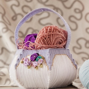 Puede incluir: Una cesta de hilo blanca y lavanda con asa, llena de ovillos de hilo rosa y morado. La cesta está decorada con adornos florales. Varios ovillos de hilo en azul, rosa y crema están junto a la cesta.