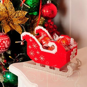 May include: A festive Christmas scene featuring a red sleigh filled with wrapped sweets, decorated with white snowflakes. The sleigh sits on a white surface, with a Christmas tree adorned with baubles and a gold poinsettia in the background.