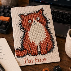 May include: A rectangular cross-stitch embroidery on light fabric featuring a fluffy, wide-eyed orange and white cat with messy fur. Below the cat, the text "I'm fine" is stitched in a dark red font. The design is centered on the fabric piece.