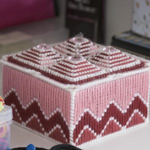 May include: A pink and burgundy needlepoint box with a geometric design, topped with four pyramid-shaped accents. Colorful bracelets and hair ties are also visible, suggesting a craft or storage theme.