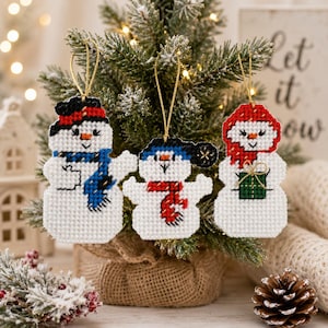 May include: Three handmade snowman decorations with gold hanging threads. Each snowman has a different coloured hat and scarf: red, blue, and black. The snowmen are displayed on a small Christmas tree. The text "Let it snow" is visible in the background.