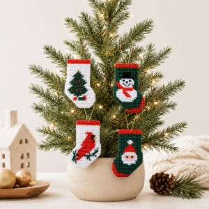 Puede incluir: Cuatro adornos navideños en miniatura con forma de calcetín colgados de un pequeño árbol de hoja perenne. Cada calcetín presenta un diseño festivo diferente: un árbol de Navidad, un muñeco de nieve, un cardenal y Papá Noel. Los calcetines son blancos y verdes con detalles rojos.