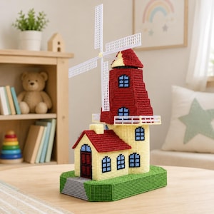 Op de afbeelding: Een kunststof handwerkobject van een windmolen met een rood dak en gele muren, voorzien van witte draaiende wieken, blauwe ramen en een donkerrode deur, gemonteerd op een groene basis met een grijs pad naar de ingang.