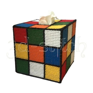 Puede incluir: Una funda de caja de pañuelos hecha a mano con forma de cubo, que se asemeja a un Cubo de Rubik. La funda está hecha con un patrón de punto de cruz en varios colores, incluyendo rojo, naranja, amarillo, verde, azul y blanco. Un pañuelo blanco es visible.