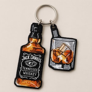 May include: A metal keychain featuring two embroidered patches: a rectangular bottle of Jack Daniel's Tennessee Whiskey with its signature black and white label, and a square glass filled with amber liquid and ice cubes, both outlined in black.