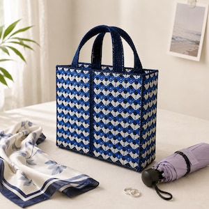 May include: A blue and white woven tote bag with navy blue handles. The bag has a geometric pattern and is on a white surface. A floral scarf, a small purple umbrella, and two silver rings are also present.