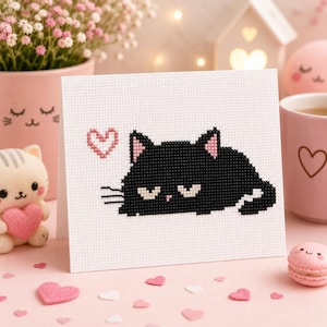 May include: A white greeting card featuring a cross-stitch design of a black cat with pink inner ears and a small pink heart above its head. The cat has a flat expression with white eyes and black whiskers, rendered in a pixelated grid pattern.