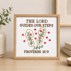 Könnte beinhalten: Ein quadratischer Holzrahmen mit einem weißen Kreuzstich-Stickbild. Der Text THE LORD GUIDES OUR STEPS und PROVERBS 16:9 ist in Braun gestickt. Dazwischen befinden sich zwei Flip-Flop-Umrisse, die mit grünen Ranken und roten Blumen gefüllt sind.