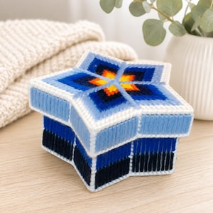 May include: A star-shaped box crafted from plastic canvas and yarn. The lid features a geometric star pattern in shades of light blue, dark blue, orange, and yellow. The sides of the box are wrapped in alternating vertical stripes of light blue, blue, and dark navy yarn.