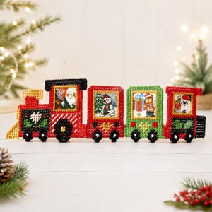 May include: A festive Christmas train decoration in red, green, and black. The carriages feature images of Santa Claus, a snowman, and a teddy bear. The train is decorated with festive details and is set against a white background.