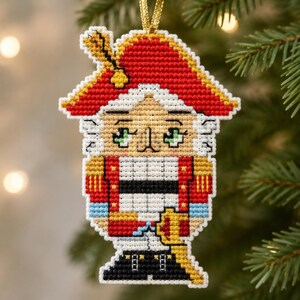 May include: A cross-stitch ornament shaped like a nutcracker soldier wearing a red hat with a gold feather, a red jacket with gold trim, light blue cuffs, and black boots. The figure has large green eyes and is suspended by a gold cord.