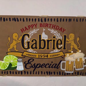 May include: A brown birthday sign with the words "Happy Birthday Gabriel 1994 Especial". The sign features illustrations of lions, limes, salt, and beer mugs. The sign is taped to a white wall.